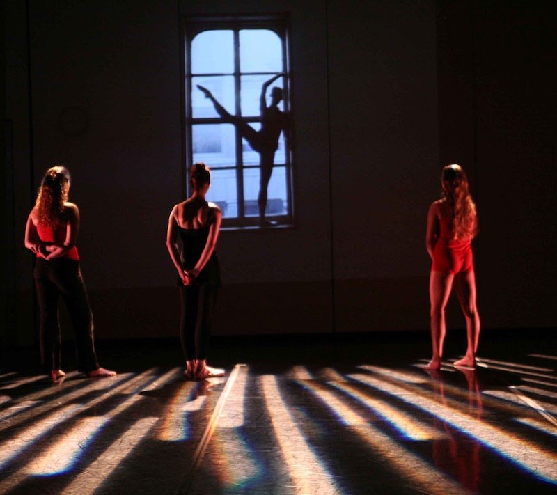 Dancers stand on stage watching a video of a dancer in silhouette extending her leg in front of a window